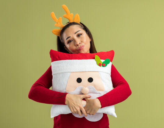 站着穿着红色圣诞毛衣的年轻女子 戴着滑稽的鹿角边 抱着圣诞枕头 站在绿色背景下 带着悲伤的表情看着相机毛衣抱着快乐