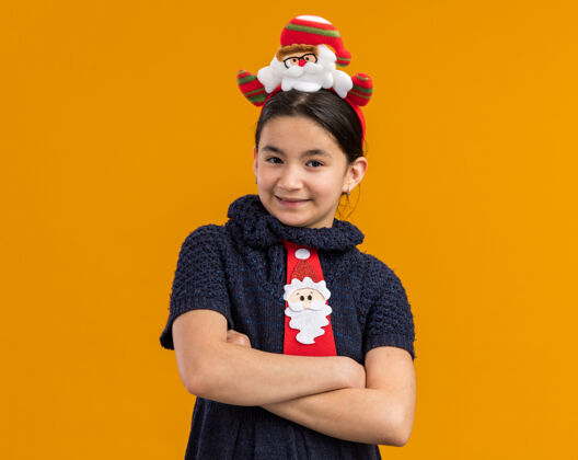 年小女孩穿着针织连衣裙 头上系着一条红色领带 头上戴着一个滑稽的圣诞戒指 双臂交叉 面带微笑快乐头新