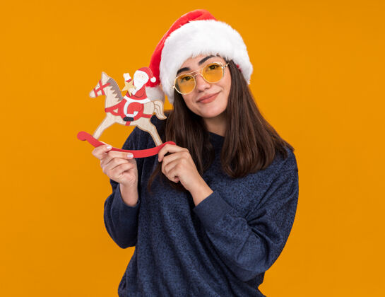 圣诞快乐戴着太阳眼镜 戴着圣诞帽的年轻高加索女孩高兴地把圣诞老人抱在摇马装饰上马年轻新