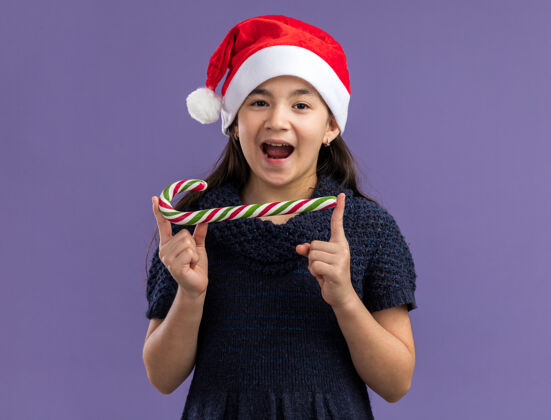 站着快乐快乐的小女孩穿着针织连衣裙戴着圣诞帽手持糖果手杖面带微笑紫色看帽子