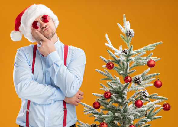 蝴蝶结一个戴着圣诞帽 戴着红眼镜 系着吊带领结的年轻人站在圣诞树旁 手放在下巴上抬起头来 想着橙色的墙年轻穿着站立