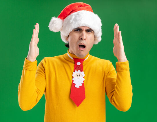 大小穿着黄色高领毛衣 戴着圣诞帽 打着滑稽领带的年轻人惊奇地用手做了一个大尺寸的手势 绿色的墙上站着测量符号高领毛衣帽子领带