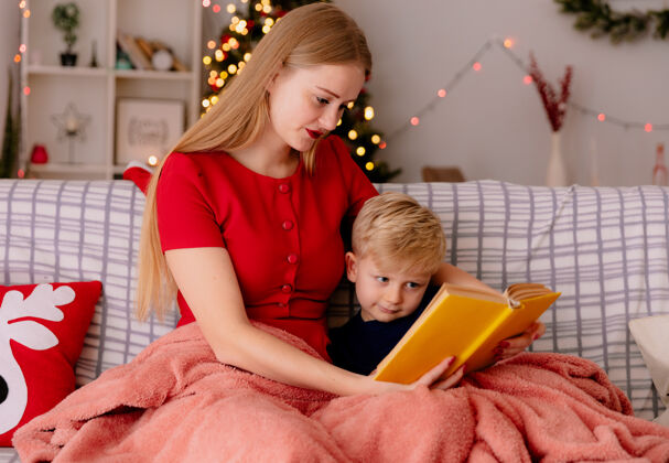 阅读快乐的妈妈穿着红色的衣服 带着她的孩子在毯子下看书 背景是圣诞树毯子圣诞树房间