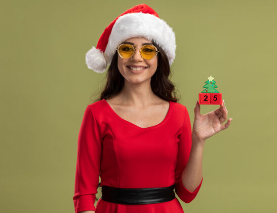 圣诞树微笑的年轻漂亮女孩戴着圣诞帽和眼镜拿着圣诞树玩具 看起来像约会快乐绿色圣诞快乐