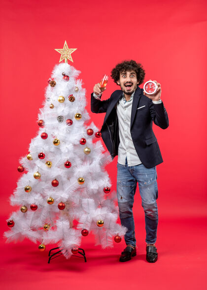 圣诞兴奋的年轻人拿着钟和一杯酒 站在红色的圣诞树旁兴奋钟站着