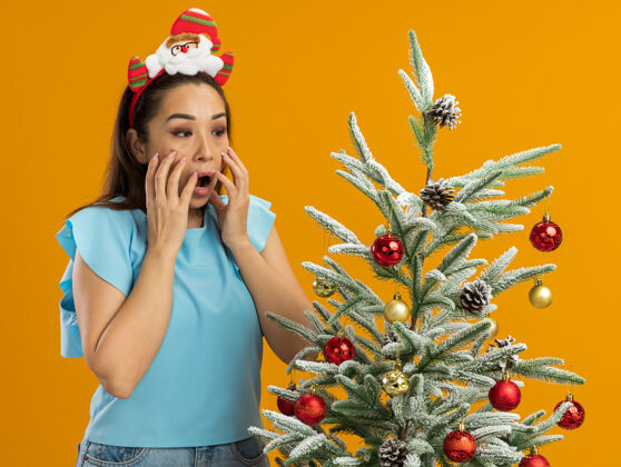 有趣身穿蓝色上衣的年轻女子头戴滑稽的圣诞环站在圣诞树旁看着它 手放在橙色背景下的脸上 震惊不已手脸年轻