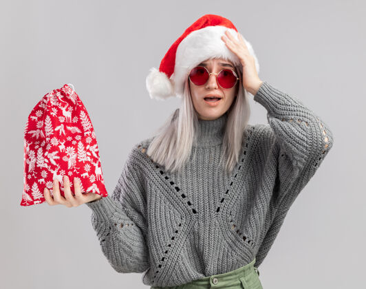 包年轻的金发女郎穿着冬天的毛衣 戴着圣诞帽 手里拿着圣诞礼物的圣诞红包 站在白色的背景下惊讶地看着镜头惊讶拿着金发