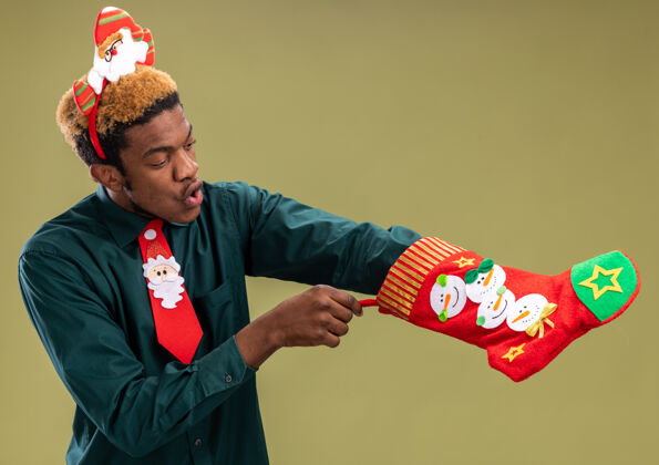 惊讶一个非洲裔美国人 戴着滑稽的圣诞帽 打着红领带 手里拿着圣诞长袜 站在绿色的墙上惊讶地看着它非洲男人长袜