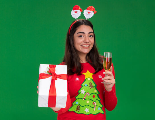 头带带着圣诞老人头带的年轻高加索女孩微笑着 手里拿着一杯香槟和一个圣诞礼盒 背景是绿色的 还有复印空间女孩微笑礼物