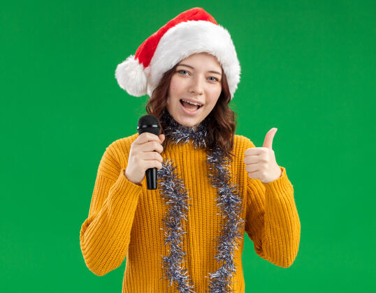 绿色戴着圣诞帽 脖子上戴着花环的年轻斯拉夫姑娘高兴地举起话筒 竖起大拇指帽子花环向上