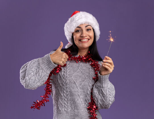 穿着快乐的年轻白人女孩戴着圣诞帽 脖子上戴着金箔花环 手里拿着节日火花灯 孤立地在紫色的墙上竖起大拇指圣诞节周围显示