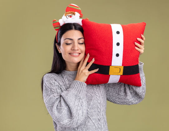 关闭微笑的年轻白人女孩戴着圣诞老人的头带 抱着圣诞老人的枕头 用它抚摸着头 闭着眼睛 在橄榄绿的背景上孤立圣诞老人头眼睛