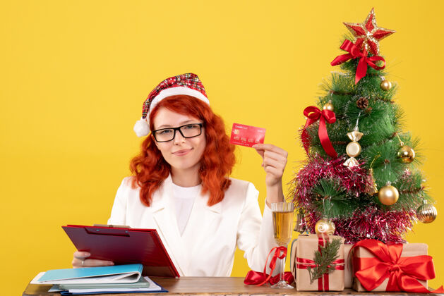 快乐正面图：女医生坐在桌子后面 手里拿着黄色背景的银行卡 还有圣诞树和礼品盒背景卡片女医生