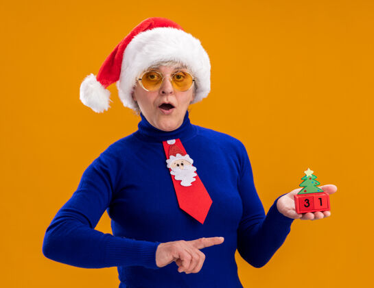 圣诞快乐一位戴着太阳眼镜 戴着圣诞帽 打着圣诞领带的老妇人拿着圣诞树的装饰物 指指点点 背景是橙色 还有复制空间老人新年领带