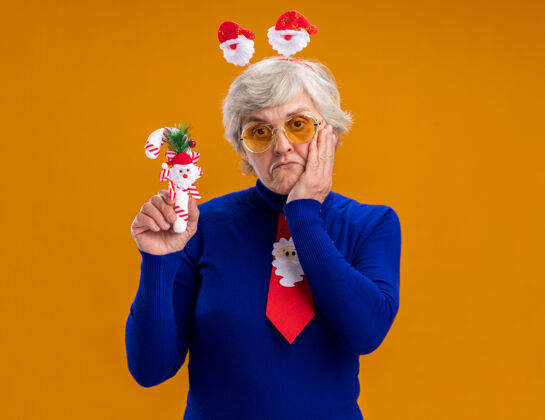 抱戴着太阳眼镜 戴着圣诞老人头带和圣诞老人领带的失望的老妇人把手放在脸上 拿着橙色背景上的糖果手杖 并留有复印空间老人领带糖果