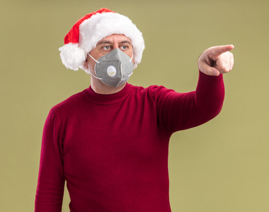 指着中年男子戴着圣诞老人帽戴着护面面具一边看一边带着忧虑的表情用食指指着旁边站在绿色背景上手指面具站着