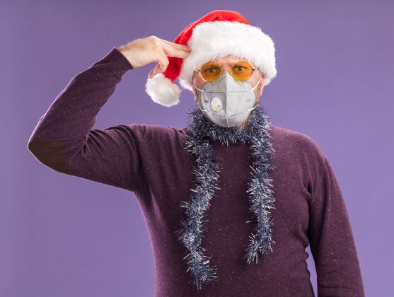 脖子受够了中年男子戴着圣诞帽 戴着防护面具 脖子上戴着金属丝花环 戴着眼镜 做着的手势 隔离在紫色的墙上圣诞节花环喂养