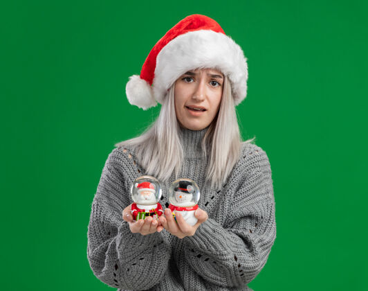 圣诞老人年轻的金发女郎穿着冬季毛衣 戴着圣诞帽 手里拿着圣诞玩具雪球 站在绿色背景下困惑地看着相机抱着困惑雪