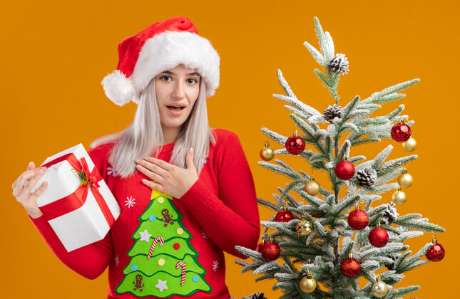 圣诞老人穿着圣诞毛衣 戴着圣诞帽 手持礼物 站在橙色背景下的圣诞树旁 看着相机 心怀感激的幸福金发女郎圣诞帽子橙色