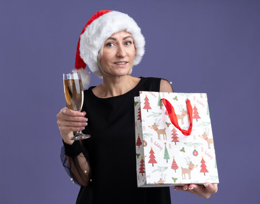 香槟微笑的中年金发女人戴着圣诞帽看着相机拿着一杯香槟和圣诞礼包孤立在紫色的背景上金发拿着相机