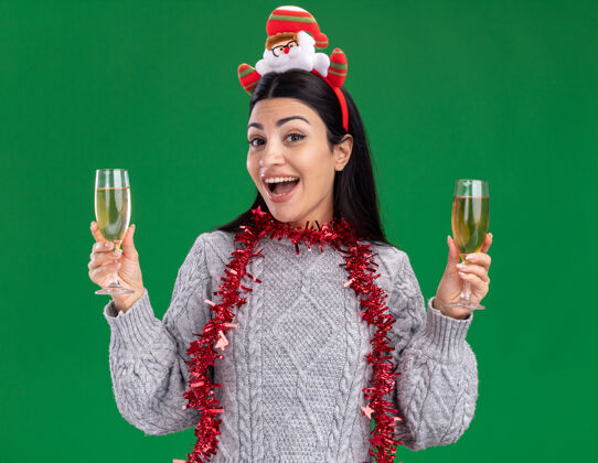 圣诞老人年轻的白人女孩戴着圣诞老人的头带 脖子上戴着金箔花环 手里拿着两杯香槟 隔着绿色的墙壁头带香槟穿着