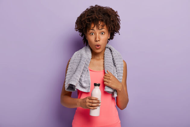 健康震惊脱水的黑皮肤女人在锻炼后喝水 穿着健身服 脖子上围着毛巾印象深刻瓶子女