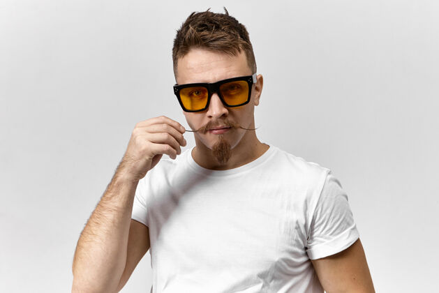 自信一幅有趣英俊的欧洲年轻男子的画像 他穿着白色t恤 戴着黄色的长方形眼镜 在电脑屏幕前工作 用发蜡卷起他时髦的小胡子脸时尚眼镜