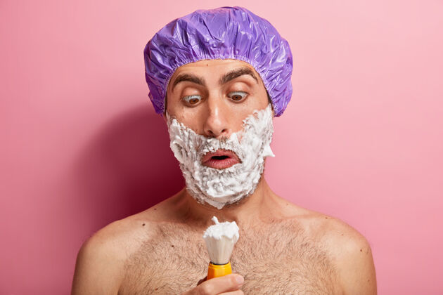 凝胶惊讶的帅哥看着刷子 贴着剃须膏 想拥有光滑的皮肤 在家里有美容程序 戴着特别的帽子男皮肤卫生