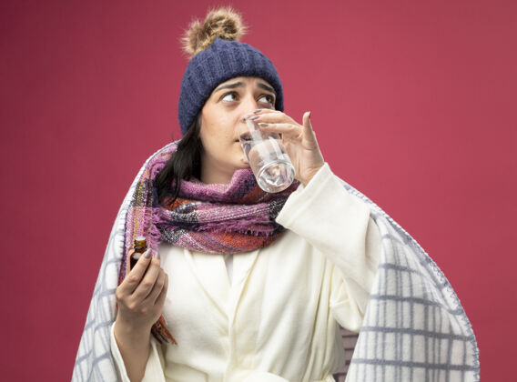 女人体弱多病的年轻白种女人穿着长袍 戴着冬帽和围巾 喝着一杯掺有药剂的水长袍帽子深红色