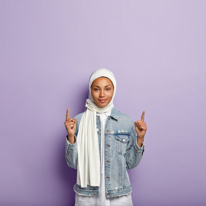 夹克深色皮肤的自信妇女的垂直镜头 指向顶部 为顾客展示了巨大的向上复制空间 戴着白色丝巾和牛仔外套 隔离在紫色的墙上客户东方头巾