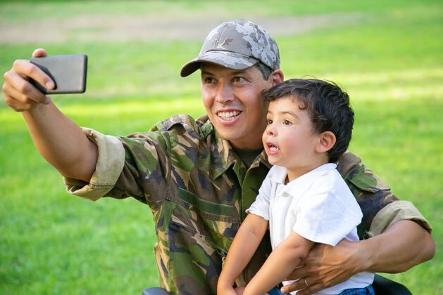 伪装快乐的残疾军人爸爸和他的小儿子一起在公园里自拍男孩坐在爸爸的膝盖上退伍军人或残疾概念士兵乐趣射击