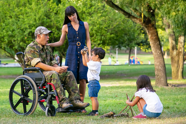 侧视图孩子们在母亲和残疾军人父亲坐在轮椅旁的公园里为营火布置木柴男孩向兴奋的父亲展示木柴残疾退伍军人或家庭户外概念男人视图完整