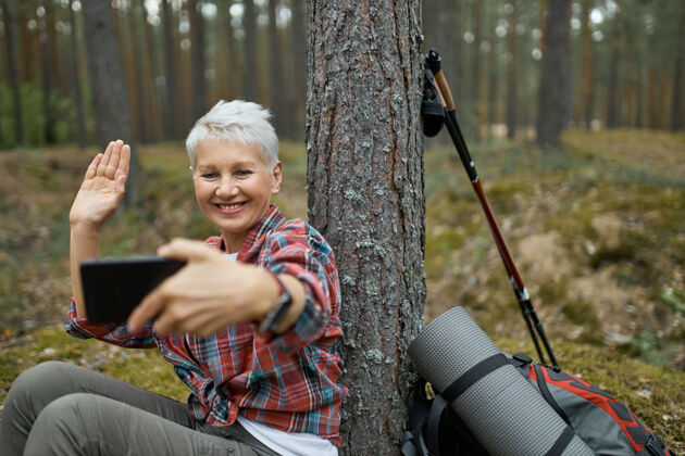 享受快乐活跃的退休女性坐在树下 带着远足装备手持手机 微笑着挥手 通过在线应用程序与朋友通过视频会议通话自然科技远足