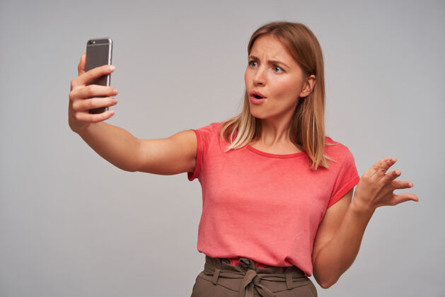 休闲室内拍摄的困惑年轻的金发女性穿着休闲服 自然化妆 一边打电话 站在白色背景下 举着智能手机身体漂亮手