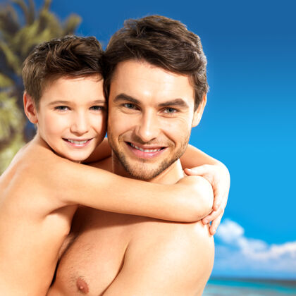 泳衣快乐微笑的父亲在热带海滩拥抱8岁的儿子的画像晒黑海洋孩子