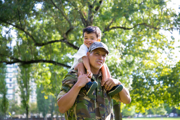 城市慈爱的父亲抱着儿子在城市公园散步快乐的白人儿子穿着制服坐在父亲的脖子上 拥抱着他 望着远方家庭团聚 做父亲 回家的概念可爱军人父亲