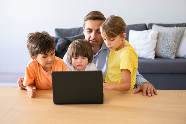 孩子白人爸爸和孩子们通过笔记本电脑看电影快乐的爸爸和可爱的孩子们坐在桌子旁可爱体贴的男孩和金发女孩看着屏幕童年和数字技术概念童年爸爸沉思