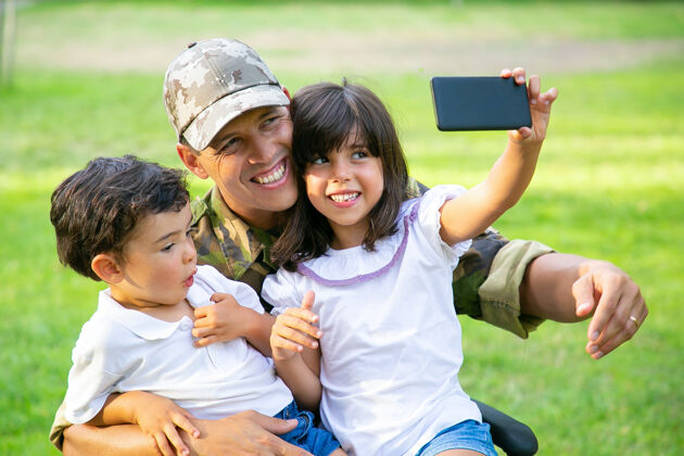 士兵两个快乐的孩子坐在爸爸的大腿上 在牢房里自拍残疾军人和孩子们在公园里散步退伍军人或残疾概念自拍男孩手机