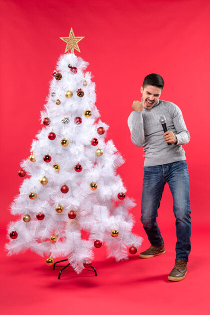 自豪圣诞节的气氛与自信的家伙穿着牛仔裤站在装饰圣诞树附近 手持麦克风自豪自信的家伙心情礼物