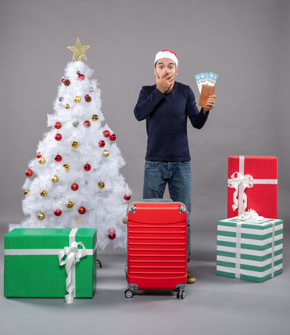 圣诞树惊讶的男人拿着他的旅行票把手放在灰色的脸上礼物包把