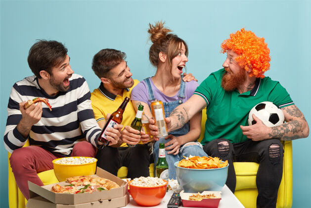 乐观三个快乐的朋友看着戴着假发的滑稽的胡须男人 碰杯啤酒 吃比萨饼 在电视上看足球比赛时玩得开心饮食四观看