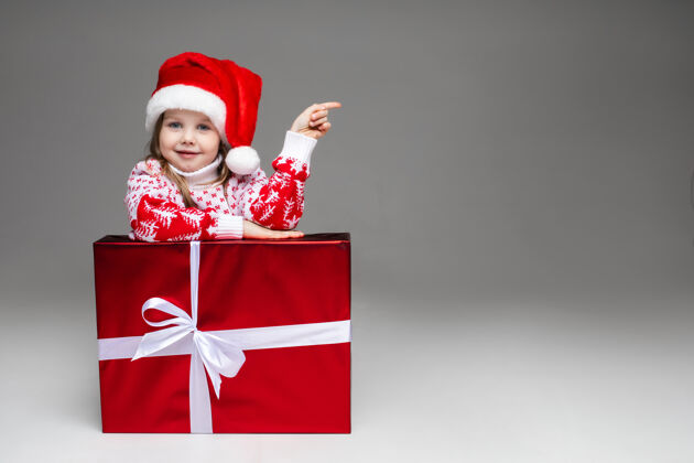 手势可爱的小女孩 穿着有图案的冬季毛衣 戴着圣诞老人帽 靠在白色蝴蝶结的包装好的圣诞礼物上可爱广告礼物
