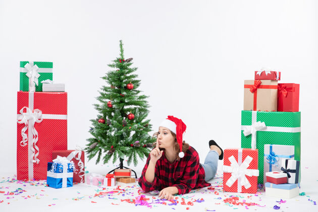 人年轻女子躺在白色墙壁上的圣诞礼物和小圣诞树周围的正视图冬青礼物礼物