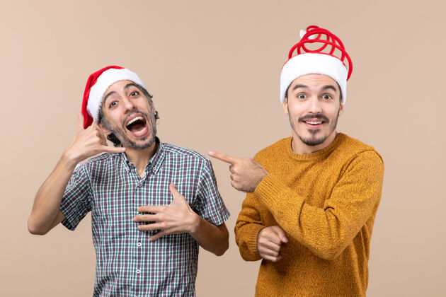 帽子正面图：两个戴着圣诞帽的微笑着的家伙 一个在打电话 另一个在米色孤立背景上用手指指着他微笑圣诞老人手指