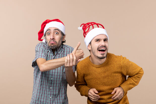 视图正面图：两个戴着圣诞帽的微笑着的家伙 一个在米色孤立背景上向另一个竖起大拇指男性两个微笑