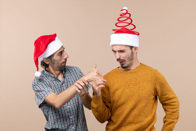 显示正面图：两个戴着圣诞帽的奇怪家伙 一个在米色孤立背景上向另一个竖起大拇指伙计们帽子前面