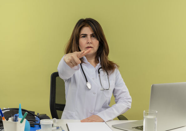 笔记本电脑严格的中年女医生 穿着医用长袍 听诊器 坐在办公桌旁 用笔记本电脑和医用工具显示你的手势和复印空间中年严格医生