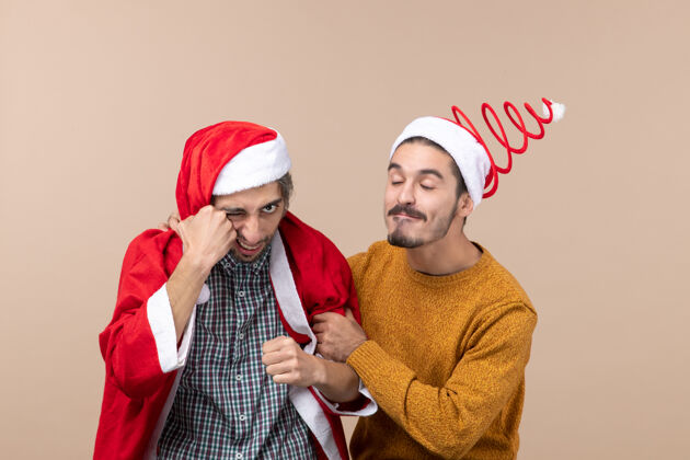 拳打正面图两个朋友 一个穿着圣诞老人外套打自己 另一个抱着朋友的手臂在米色孤立的背景上两个朋友圣诞老人外套