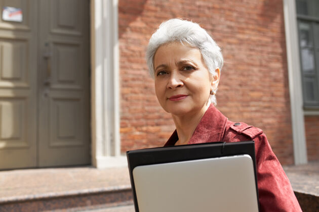 小工具现代忙碌的白发中年妇女在砖房外摆姿势 带着时尚的数码设备进行远程工作或在线教育便携连接电脑