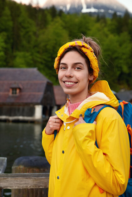 白天可爱开朗的女士 带着愉快的微笑 穿着带兜帽的黄色雨衣 背着背包头带乐观刮风